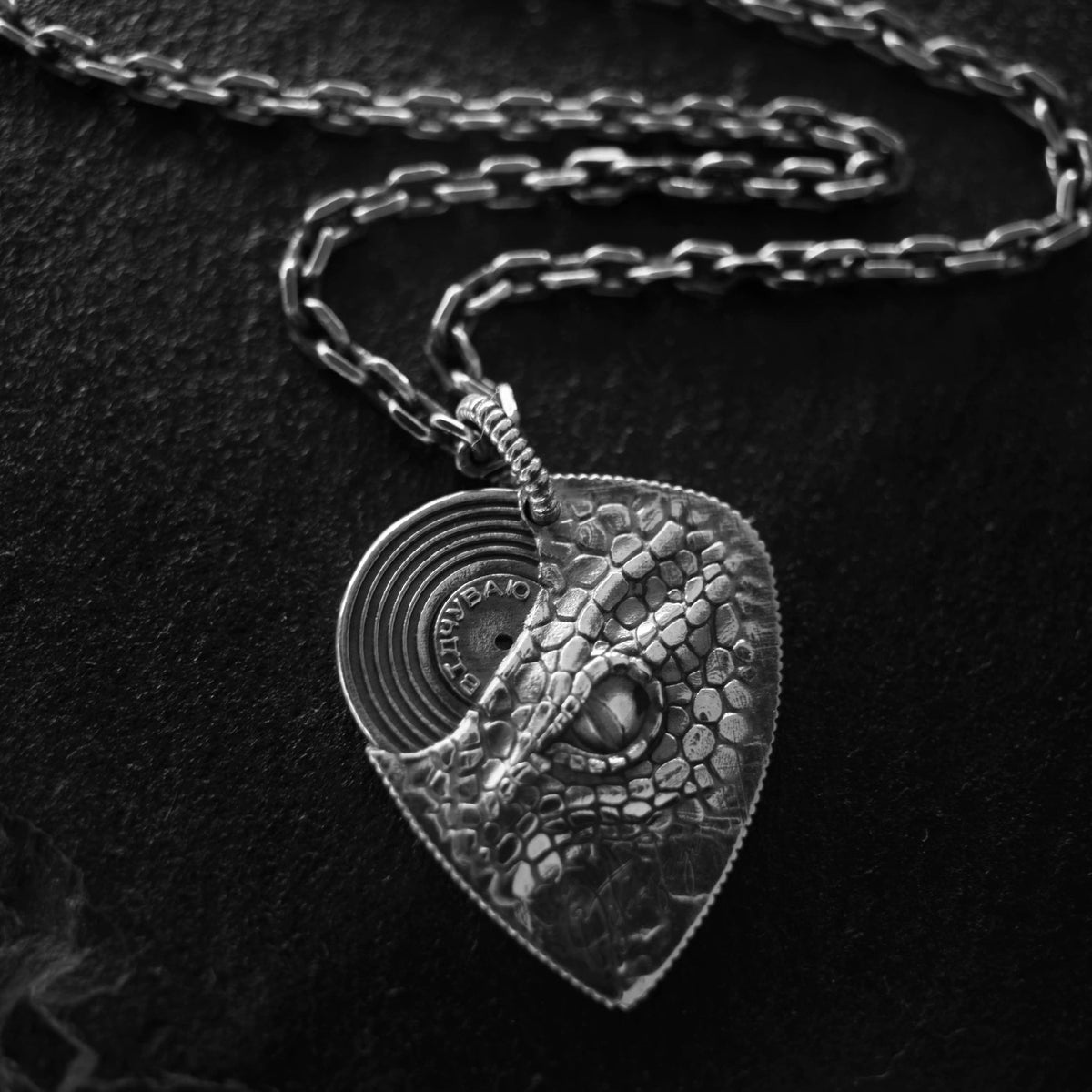 A silver-toned pendant necklace featuring a guitar pick design with a crocodile or dragon face motif and a circular inset, displayed on a dark textured surface.