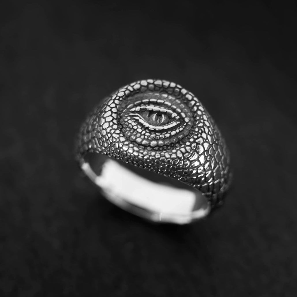 A textured silver ring featuring a raised eye motif of rattlesnake on a dark, textured background.