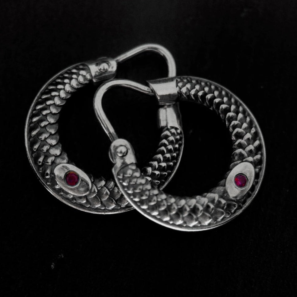 A pair of silver-toned hoop earrings with a textured, snake-like pattern and small red gemstone accents, displayed on a dark textured background.