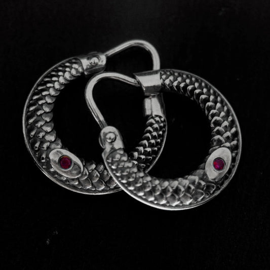 A pair of silver-toned hoop earrings with a textured, snake-like pattern and small red gemstone accents, displayed on a dark textured background.