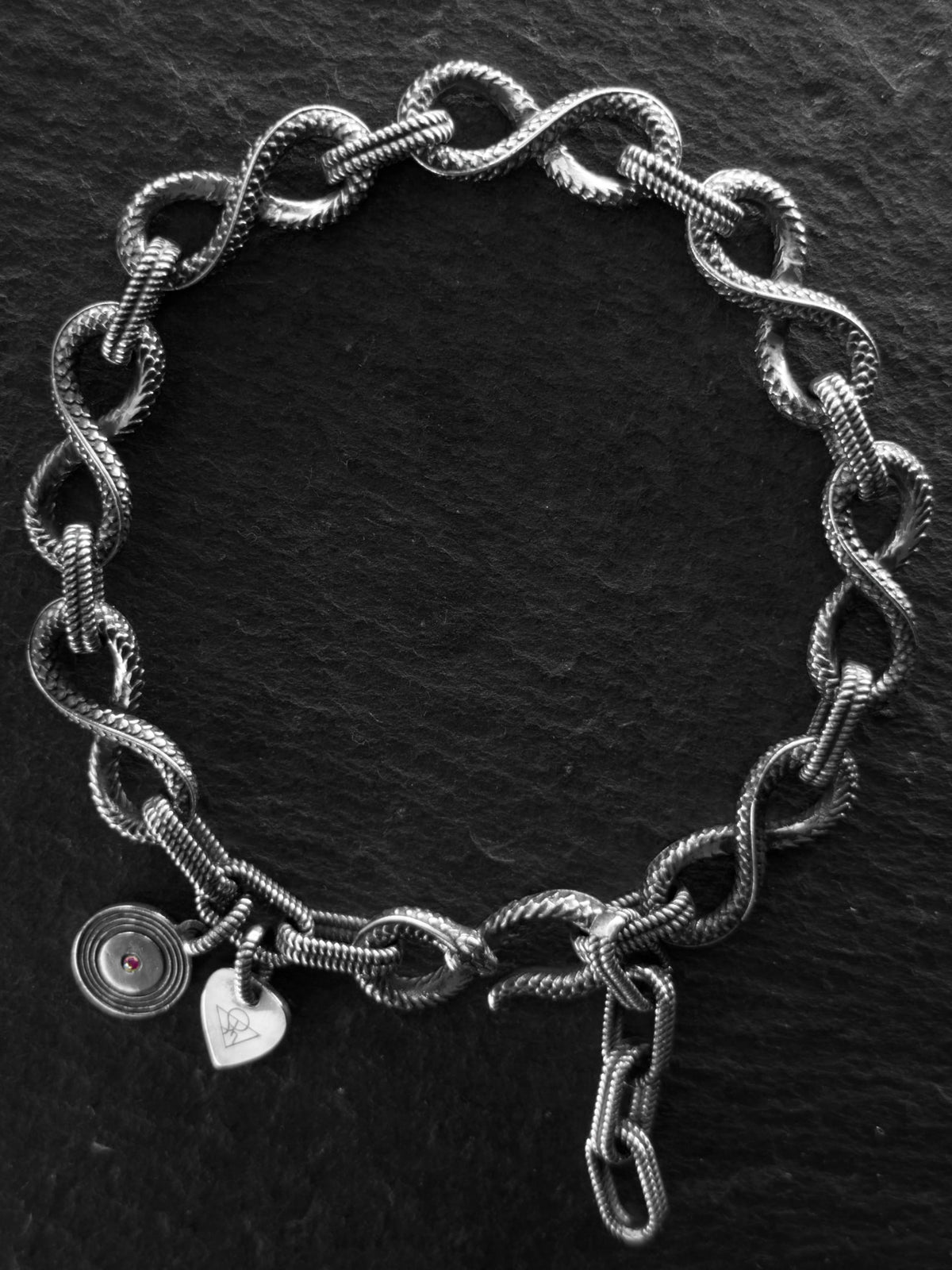 A silver-toned chain bracelet with textured interlocking links, featuring two small charms—a round disc charm and a heart-shaped charm—resting on a dark slate surface.